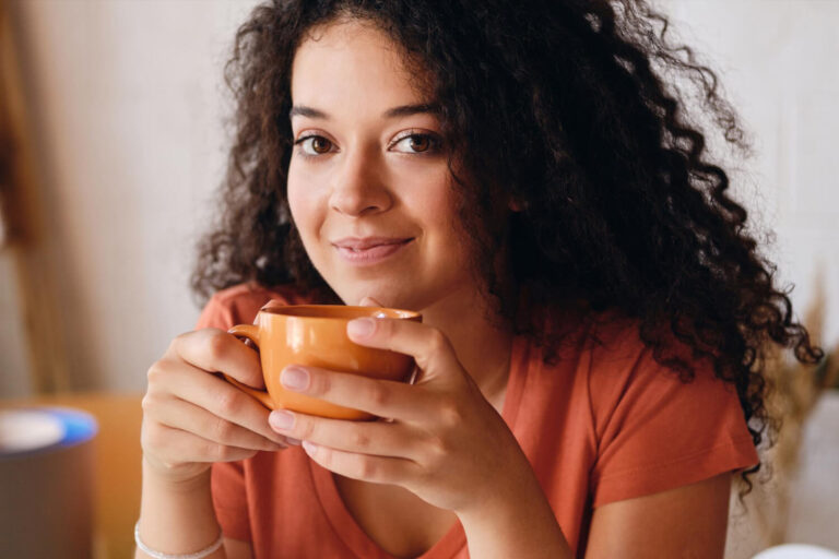 Mulher de cabelos cacheados segurando uma xícara, refletindo se lentes de porcelana mancham com café na Mooca na AGN Odontologia.
